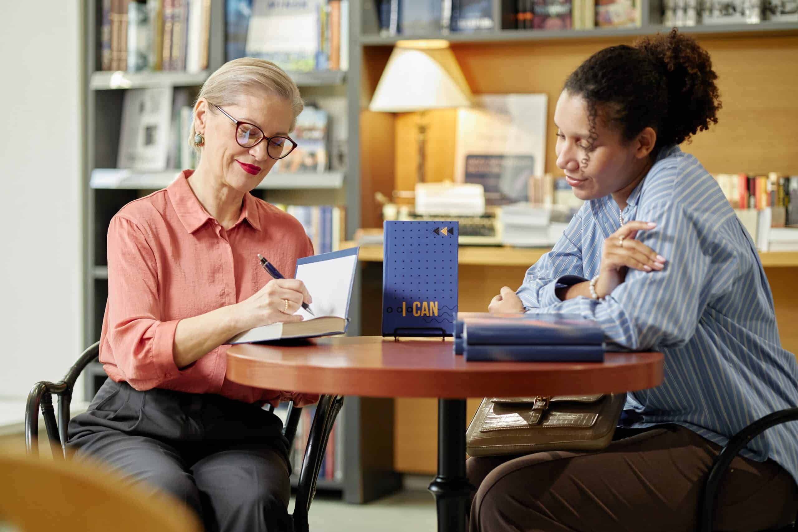 Author writing in book with a woman glancing over sitting at a table