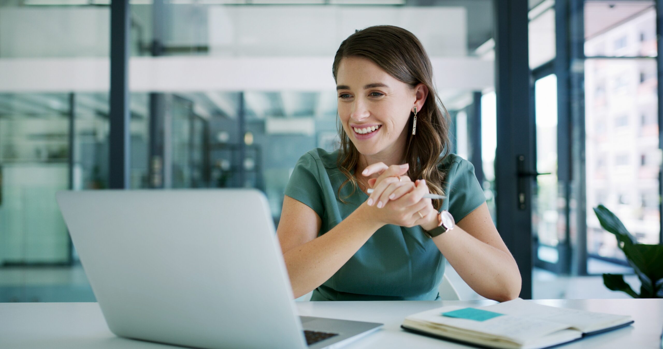Woman designer speaking on a laptop with notebook of notes laid out on desk.