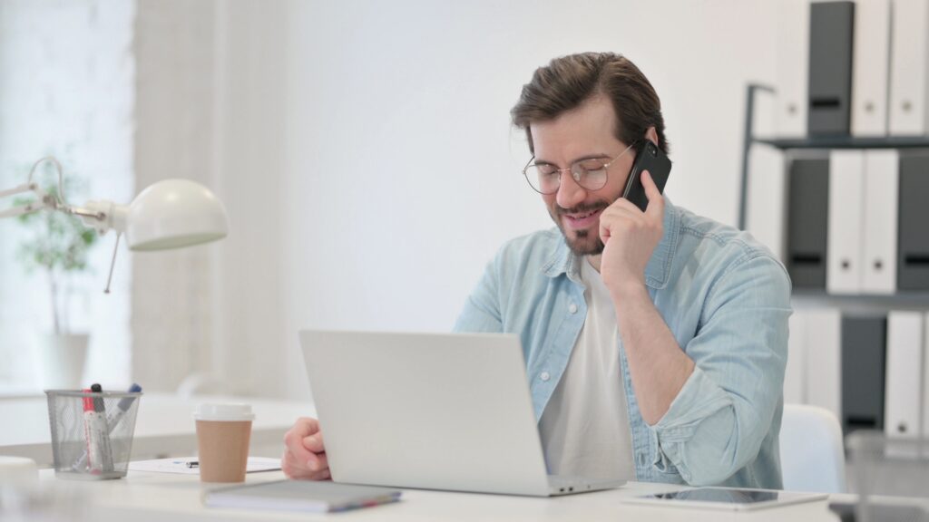 Man sitting at a desk holding a phone with a laptop in front.