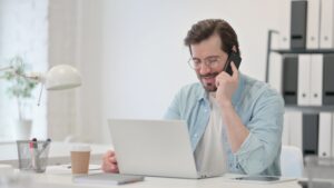 Man sitting at a desk holding a phone with a laptop in front.