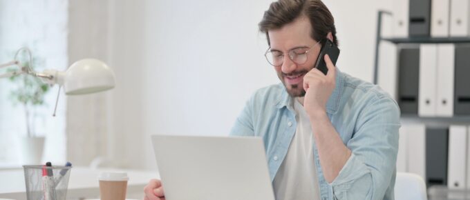 Man sitting at a desk holding a phone with a laptop in front.