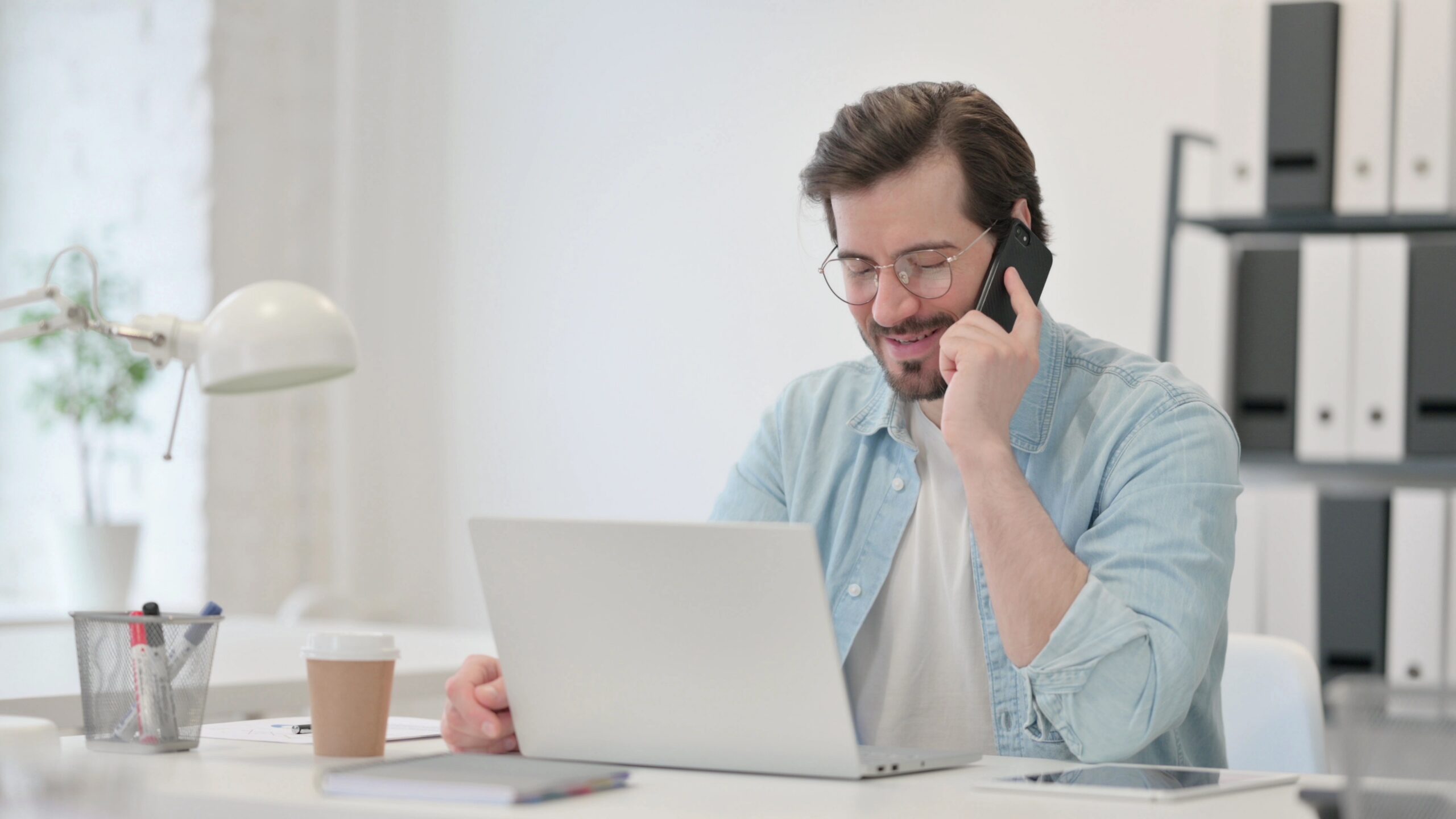Man sitting at a desk holding a phone with a laptop in front.