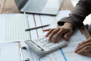 Woman typing on the calculator. Statistical data, forms, and laptop on desk.